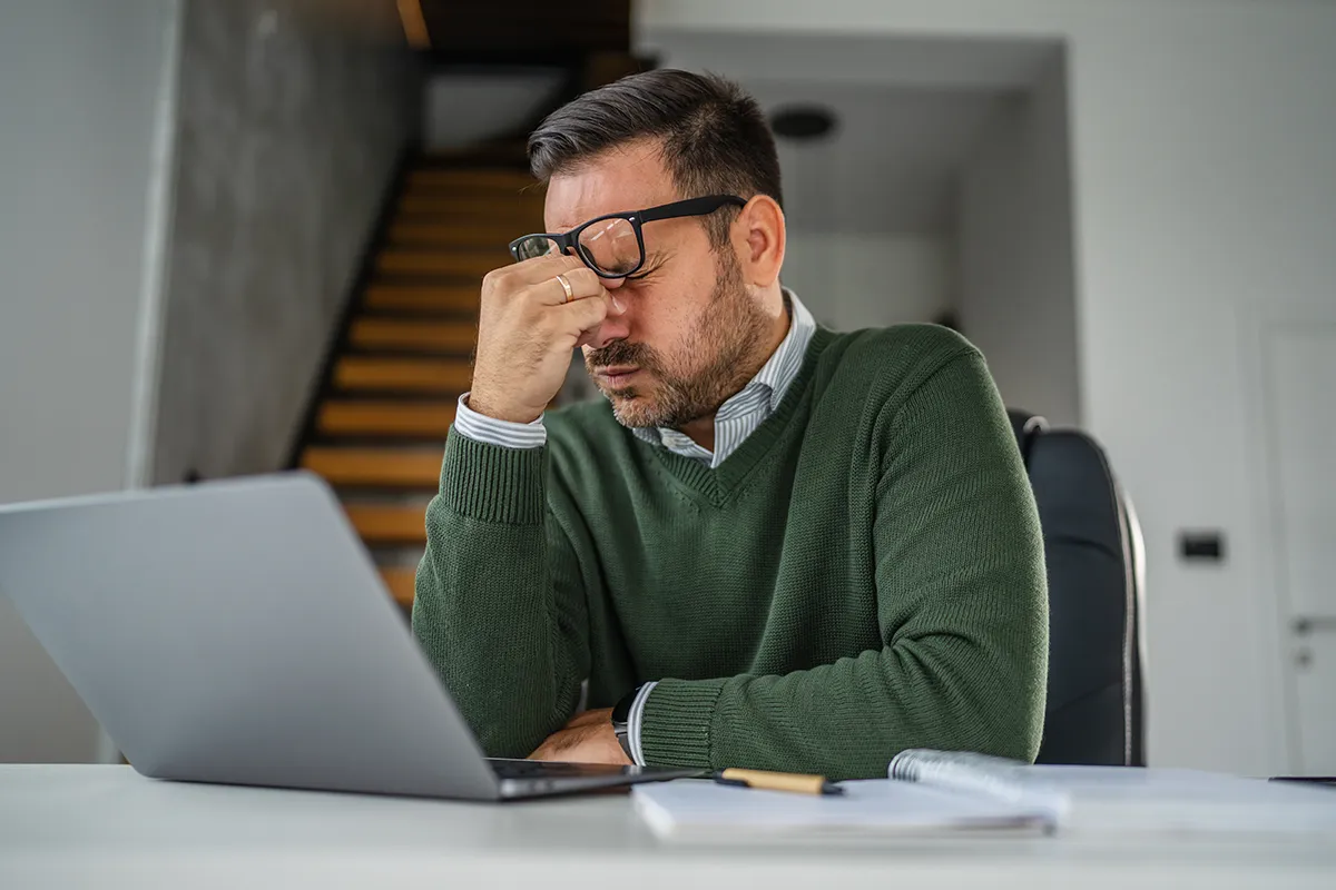 a man massaging the bridge of his nose while working on a laptop at home